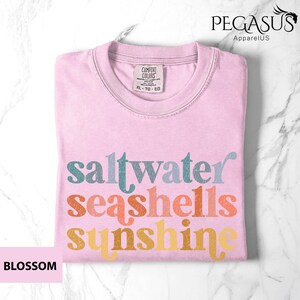May include: A light pink t-shirt with the words "saltwater seashells sunshine" in teal, orange, and yellow. The shirt is folded and has a tag that says "Comfort Colors". The word "Blossom" is in a pink box.
