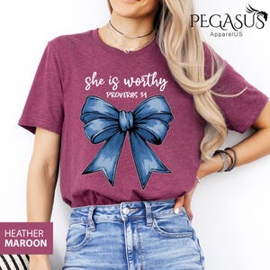 May include: Heather maroon t-shirt with a blue bow and the text "She is worthy Proverbs 31" printed on the front.
