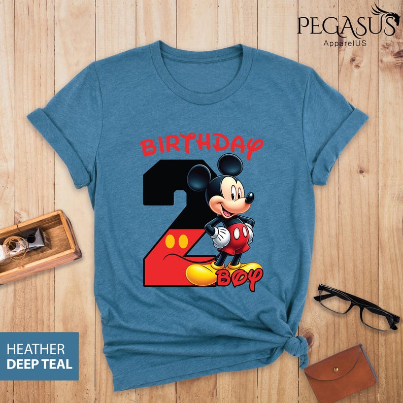 May include: A teal blue t-shirt featuring a graphic of Mickey Mouse with the number 2, the word "BIRTHDAY" in red, and the word "BOY" in yellow. The shirt is made of a soft material and is suitable for a child's birthday.