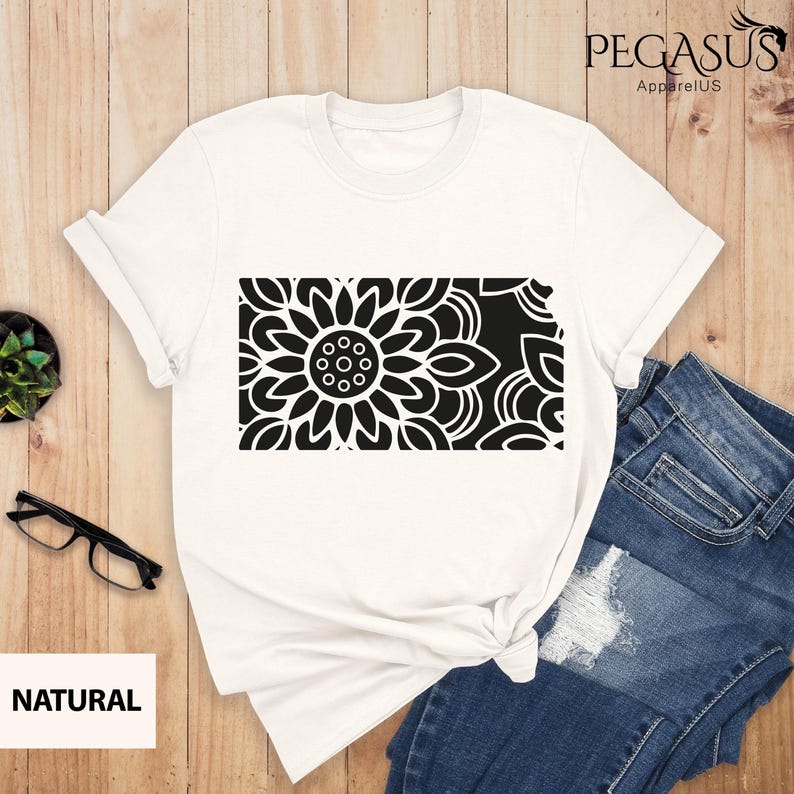 May include: A white t-shirt with a black mandala design. The design is a stylized flower with intricate details. The t-shirt is laid out on a wooden surface with a pair of jeans and a plant in the background.