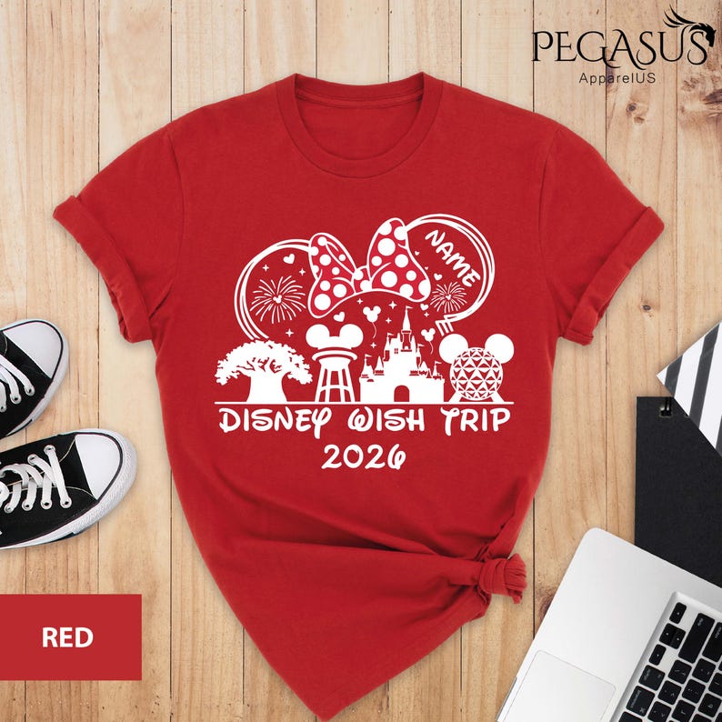May include: Red t-shirt with a white graphic featuring a Minnie Mouse bow, fireworks, and a castle. The text "DISNEY WISH TRIP 2026" is printed below the graphic. The shirt is displayed on a wooden surface.