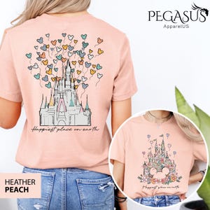 Op de afbeelding: Een roze T-shirt met een afbeelding van Assepoester's kasteel en Mickey Mouse ballonnen. De tekst "Happiest place on earth" is onder het kasteel gedrukt. Het shirt is verkrijgbaar in Heather Peach.