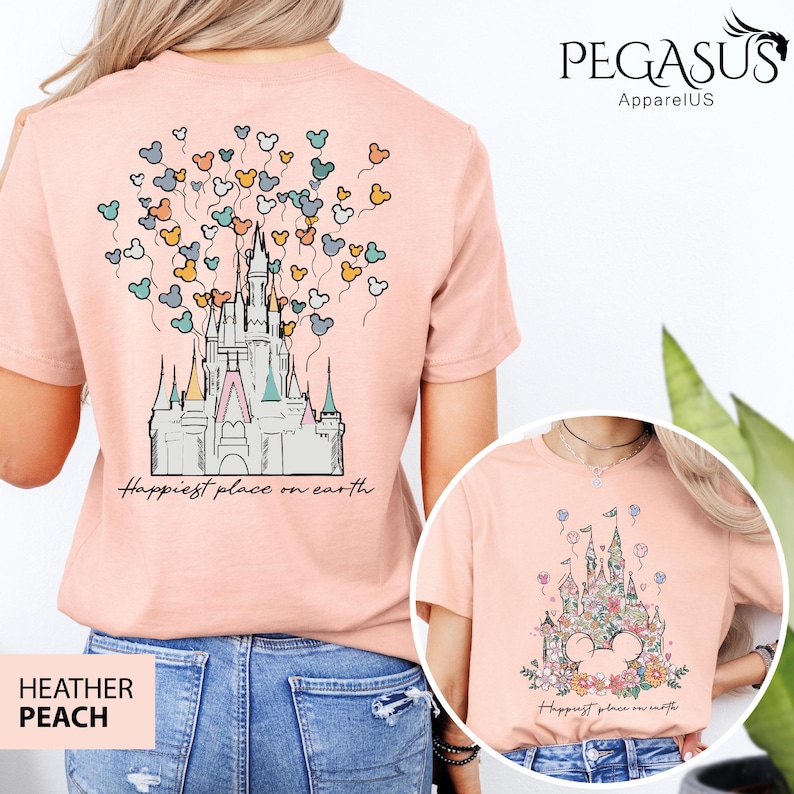 Op de afbeelding: Een roze T-shirt met een afbeelding van Assepoester's kasteel en Mickey Mouse ballonnen. De tekst "Happiest place on earth" is onder het kasteel gedrukt. Het shirt is verkrijgbaar in Heather Peach.