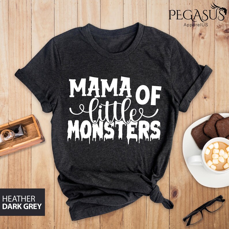 May include: Dark heather grey t-shirt with the text "MAMA OF little MONSTERS" in white. The shirt is folded on a wooden surface. The words "HEATHER DARK GREY" are printed on the bottom left.