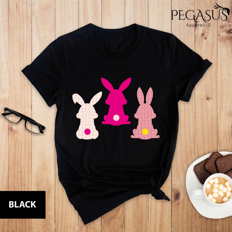May include: Black t-shirt featuring three Easter bunny silhouettes in pink and white. The bunnies have textured patterns and a yellow or white tail. The text "BLACK" is at the bottom left of the image, and "PEGASUS ApparelUS" is at the top right.