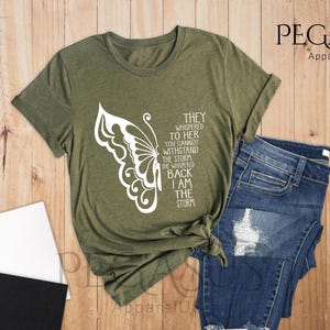 Op de afbeelding: Een groen T-shirt met een witte vlinderprint en de tekst "They whispered to her you cannot withstand the storm she whispered back I am the storm".