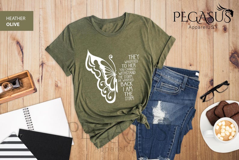 Op de afbeelding: Een groen T-shirt met een witte vlinderprint en de tekst "They whispered to her you cannot withstand the storm she whispered back I am the storm".