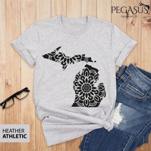 May include: A heather grey t-shirt with a black mandala design of the state of Michigan. The shirt is laid on a wooden surface with a pair of glasses and a pair of jeans.