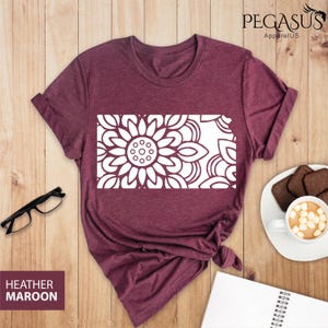 May include: A heather maroon t-shirt with a white mandala design in the shape of the state of Kansas. The t-shirt is laid flat on a wooden surface.