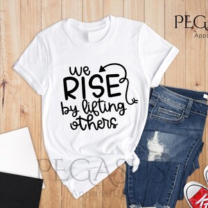 Pu&ograve; includere: Maglietta bianca con testo nero che recita "We Rise by lifting others" con una freccia che punta verso l'alto. La maglietta &egrave; appoggiata su una superficie di legno con un paio di jeans blu e scarpe da ginnastica rosse.
