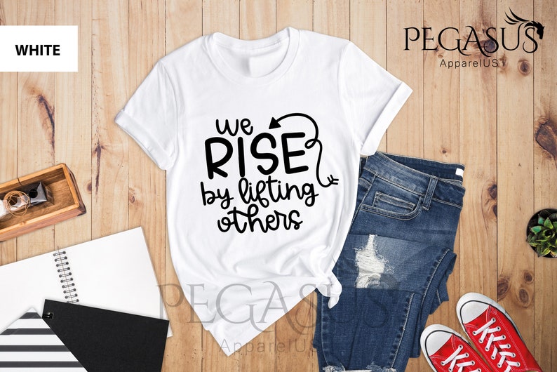 Pu&ograve; includere: Maglietta bianca con testo nero che recita "We Rise by lifting others" con una freccia che punta verso l'alto. La maglietta &egrave; appoggiata su una superficie di legno con un paio di jeans blu e scarpe da ginnastica rosse.