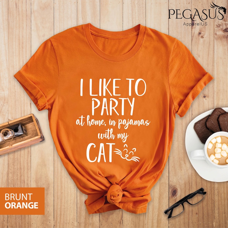 Op de afbeelding: Oranje T-shirt met de tekst "Ik vind het leuk om te feesten thuis, in pyjama met mijn kat" in witte letters. De kat heeft een witte omtrek en een wit gezicht met twee witte snorharen.