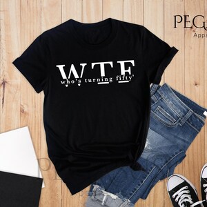 May include: Black t-shirt with white text that reads "WTF who's turning fifty?"