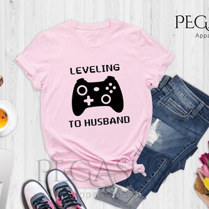 May include: Pink t-shirt with a black video game controller graphic and the text "LEVELING TO HUSBAND".