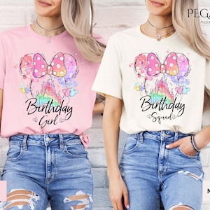 Può includere: Due magliette, una rosa e una naturale, con una grafica colorata di un fiocco, un castello e le parole "Birthday Girl" e "Birthday Squad". Le magliette hanno maniche corte e sono abbinate a jeans blu.