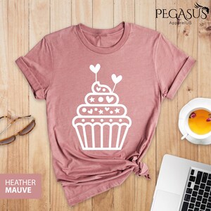 May include: A heather mauve t-shirt with a white cupcake graphic. The cupcake has white frosting with hearts and stars on top. The cupcake is topped with two white hearts on sticks.
