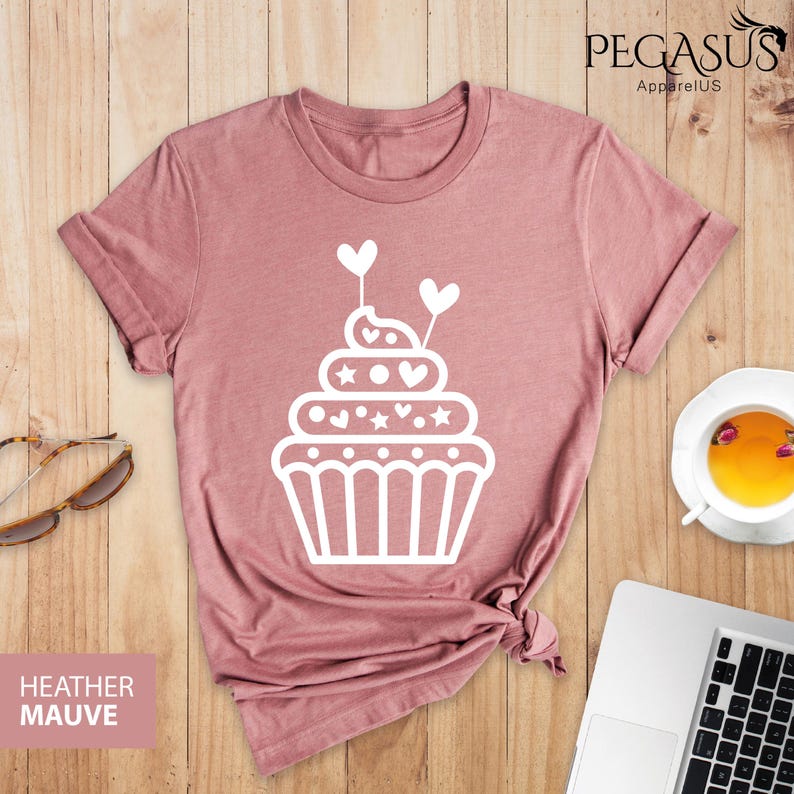 May include: A heather mauve t-shirt with a white cupcake graphic. The cupcake has white frosting with hearts and stars on top. The cupcake is topped with two white hearts on sticks.