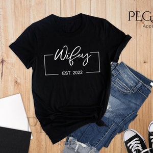 May include: Black t-shirt with white text that says "Wifey Est. 2022" in a box.