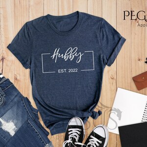 May include: A heather navy blue t-shirt with a white graphic that says "Hubby Est. 2022" in a rectangular frame.