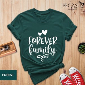 Pode incluir: Uma camiseta verde escura com o texto "FOREVER family" em letras brancas. O texto est&aacute; em uma fonte cursiva e &eacute; acompanhado por tr&ecirc;s cora&ccedil;&otilde;es brancos.