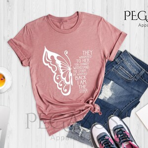 Op de afbeelding: Een heather mauve t-shirt met een witte vlinder afbeelding en de tekst "They whispered to her you cannot withstand the storm, she whispered back I am the storm".