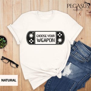 Può includere: Maglietta bianca con una grafica nera di una console per videogiochi con il testo "Choose Your Weapon".