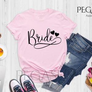 May include: Pink t-shirt with the word "Bride" in black script with two small black hearts above the word. The shirt is laid out on a white wooden surface with a pair of sunglasses, a cup of tea, a laptop, a pair of sneakers, a pair of denim shorts, and a bouquet of flowers.