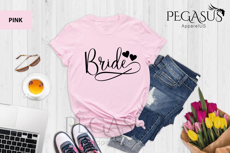 May include: Pink t-shirt with the word "Bride" in black script with two small black hearts above the word. The shirt is laid out on a white wooden surface with a pair of sunglasses, a cup of tea, a laptop, a pair of sneakers, a pair of denim shorts, and a bouquet of flowers.
