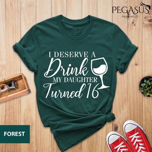 May include: A dark green t-shirt with white text that reads "I deserve a drink my daughter turned 16" and a white wine glass graphic.