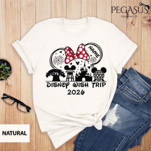 May include: White t-shirt featuring a Minnie Mouse bow with red polka dots, fireworks, and a castle graphic. The text "Disney Wish Trip 2026" is printed below the design. The shirt is paired with blue jeans.