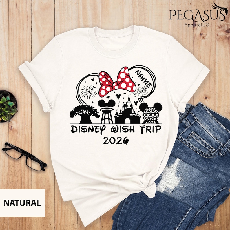 May include: White t-shirt featuring a Minnie Mouse bow with red polka dots, fireworks, and a castle graphic. The text "Disney Wish Trip 2026" is printed below the design. The shirt is paired with blue jeans.