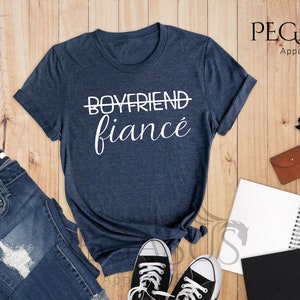 May include: A heather navy blue t-shirt with the text "BOYFRIEND" crossed out and "fianc&eacute;" written in cursive below it. The shirt is laid flat on a wooden surface.