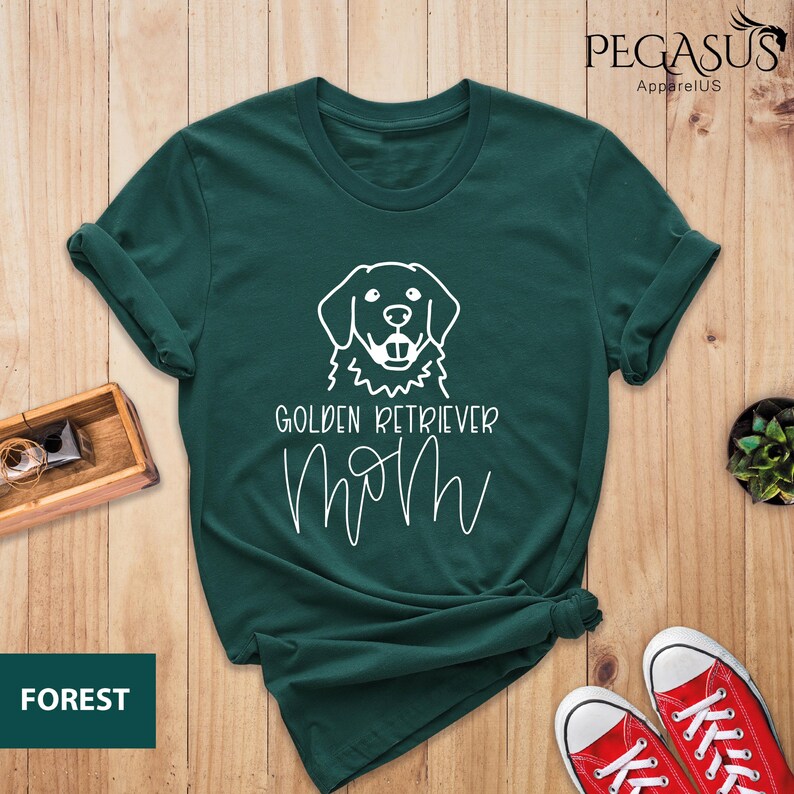 Op de afbeelding: Een donkergroen T-shirt met een witte omtrek van een golden retriever hond en de tekst "Golden Retriever Mom" in wit.