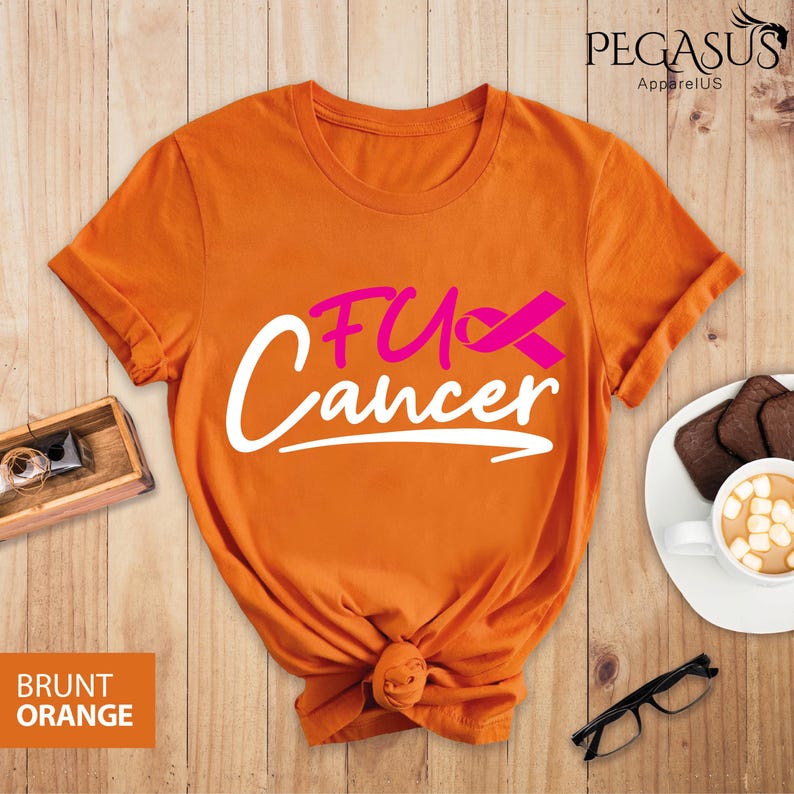 May include: Orange t-shirt with the text "F*ck Cancer" in white with a pink ribbon. The shirt is folded and displayed on a wooden surface.