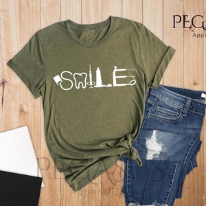 May include: A heather olive t-shirt with a white graphic of dental tools spelling out the word "SMILE". The t-shirt is laid out on a wooden surface with a pair of jeans, a notebook, and a cup of coffee.