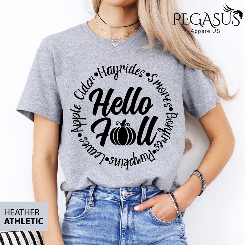 May include: Heather gray athletic t-shirt with the text "Hello Fall" in a circular design, including a pumpkin graphic. The design also features words like "Apple," "Leaves," and "Pumpkins."