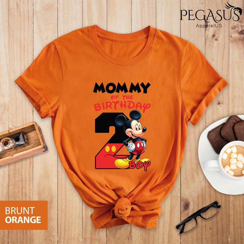 May include: Burnt orange t-shirt featuring a graphic of Mickey Mouse with the text "MOMMY OF THE BIRTHDAY" and the number "2". The shirt also has the word "BOY" below Mickey Mouse. The shirt is knotted at the bottom.