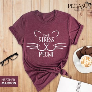 Op de afbeelding: Heather maroon t-shirt met een witte kattenkop-afbeelding en de tekst "Don't Stress Meowt".