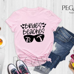 Pu&ograve; includere: Maglietta rosa con la scritta "Bride's Beaches" e un paio di occhiali da sole con palme riflesse nelle lenti.