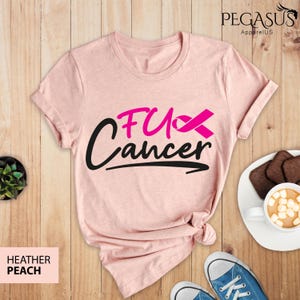 May include: Pink heather peach t-shirt with the text "F*ck Cancer" in black with a pink ribbon. The shirt is laid flat on a wooden surface.