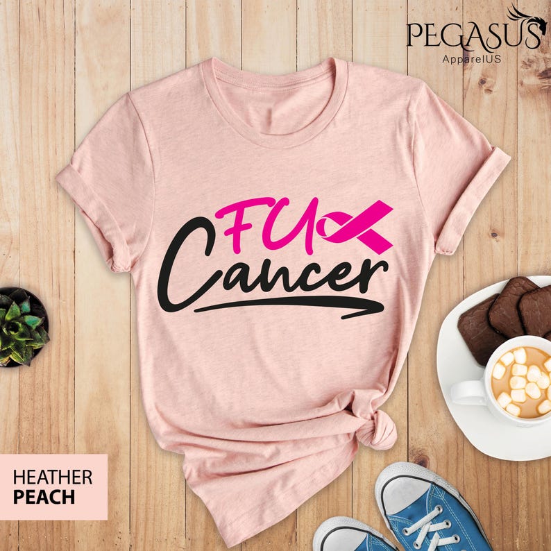 May include: Pink heather peach t-shirt with the text "F*ck Cancer" in black with a pink ribbon. The shirt is laid flat on a wooden surface.