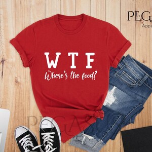 May include: Red t-shirt with white text that reads "WTF Where's the food?" The shirt is laid out on a wooden surface with a pair of black trainers and a pair of blue denim shorts.