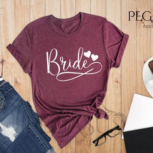 May include: A heather maroon t-shirt with the word "Bride" in white script with two white hearts above the word.