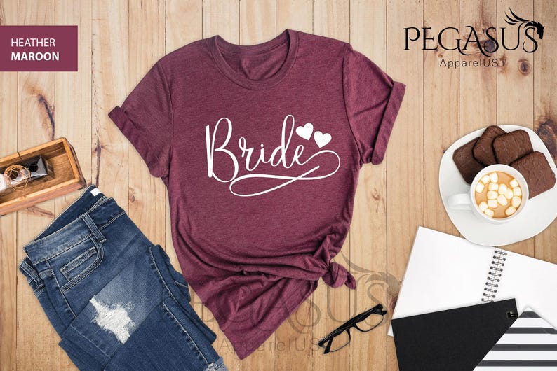 May include: A heather maroon t-shirt with the word "Bride" in white script with two white hearts above the word.