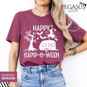 May include: Heather maroon t-shirt with a white graphic that says "HAPPY CAMP-O-WEEN". The design includes a tree, bats, a witch on a broom, and a camper. The shirt is a crew neck.