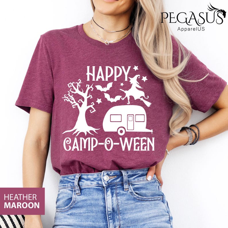 May include: Heather maroon t-shirt with a white graphic that says "HAPPY CAMP-O-WEEN". The design includes a tree, bats, a witch on a broom, and a camper. The shirt is a crew neck.