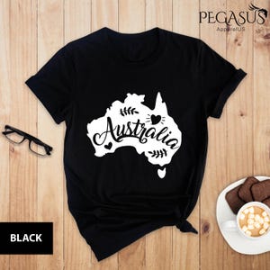 Op de afbeelding: Zwart T-shirt met een witte omtrek van het continent Australi&euml; en het woord "Australi&euml;" in een gestileerd lettertype. Er zijn kleine hartjes rond de omtrek.