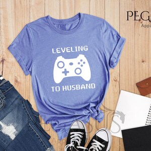 May include: A light blue t-shirt with the text "LEVELING TO HUSBAND" in white pixelated font. A white video game controller graphic is above the text.