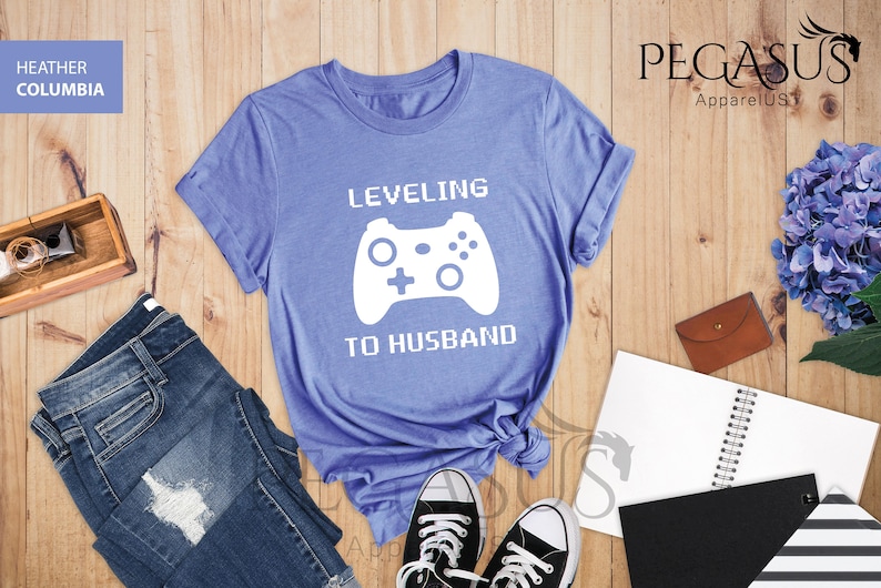 May include: A light blue t-shirt with the text "LEVELING TO HUSBAND" in white pixelated font. A white video game controller graphic is above the text.
