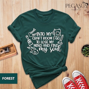 K&ouml;nnte beinhalten: Ein dunkelgr&uuml;nes T-Shirt mit wei&szlig;em Text, der "Into my craft room I go to lose my mind and find my soul" lautet. Der Text ist von Bildern von Bastelwerkzeugen umgeben, darunter ein Pinsel, eine N&auml;hmaschine und eine S&auml;ge.